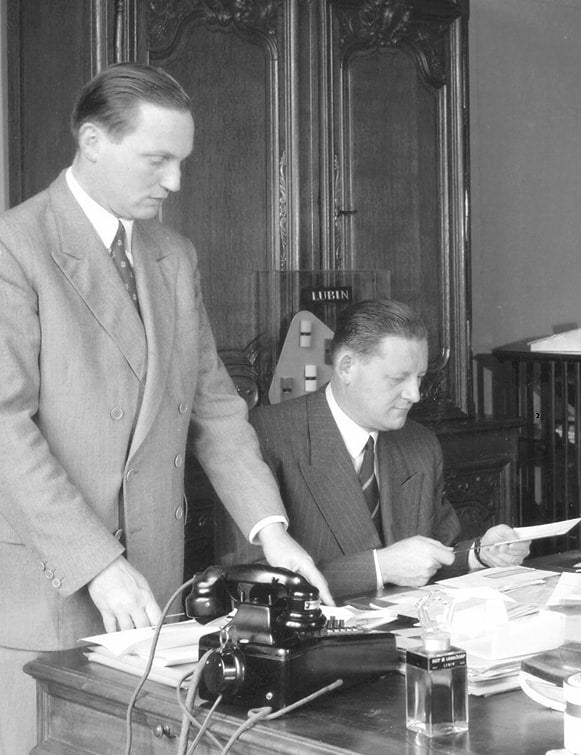 Photographie en noir et blanc de deux hommes en costume dans un bureau lambrissé : l'un debout, l'autre assis à une table de travail encombrée de papiers, près d'un téléphone noir.