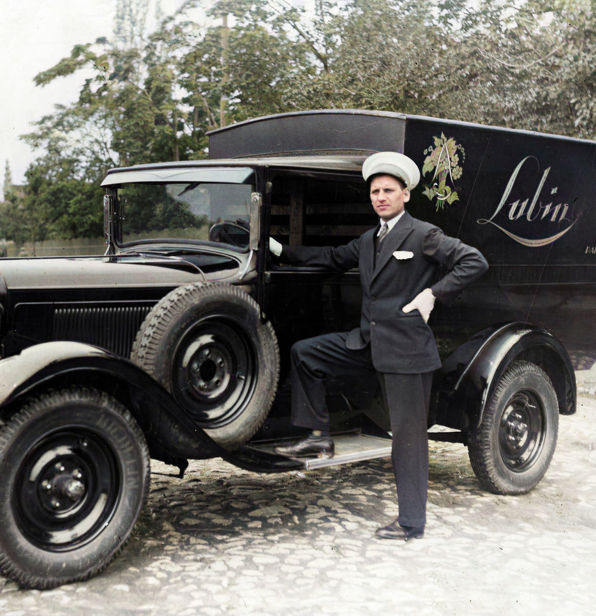 Photographie ancienne colorisée : un livreur en costume sombre et casquette blanche, une main sur la hanche, pose devant une camionnette de livraison noire dont le flanc porte le nom Lubin peint en lettres cursives surmonté d'un petit bouquet fleuri, sur un sol pavé bordé d'arbres.