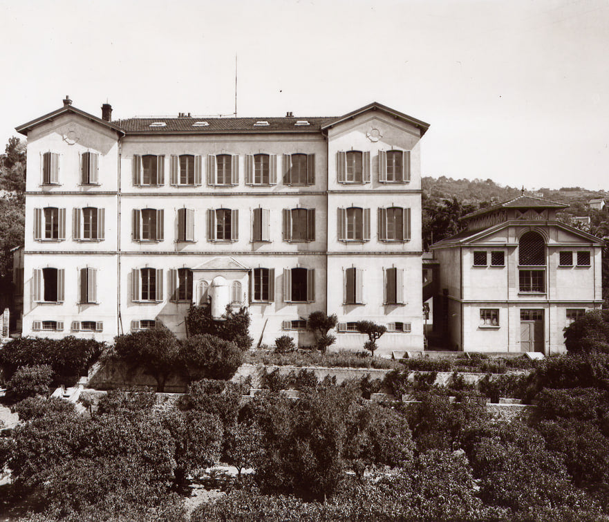 Photographie ancienne en sépia d'un grand bâtiment blanc de trois étages aux nombreuses fenêtres à volets, surmonté d'un mât, entouré d'un jardin aux allées dessinées, avec des collines à l'arrière-plan.
