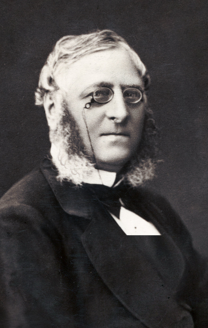 Portrait photographique ancien, en buste, d'un homme d'âge mûr aux favoris et à la barbe fournis, portant un monocle, vêtu d'une redingote sombre sur une chemise blanche à large nœud.