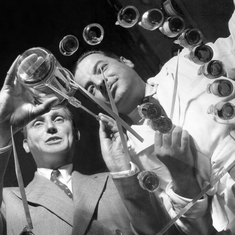 Photographie en noir et blanc prise en contre-plongée : deux hommes, l'un en blouse blanche, l'autre en costume, entourés de flacons d'essences et de longues mouillettes, dans un laboratoire de parfumeur.