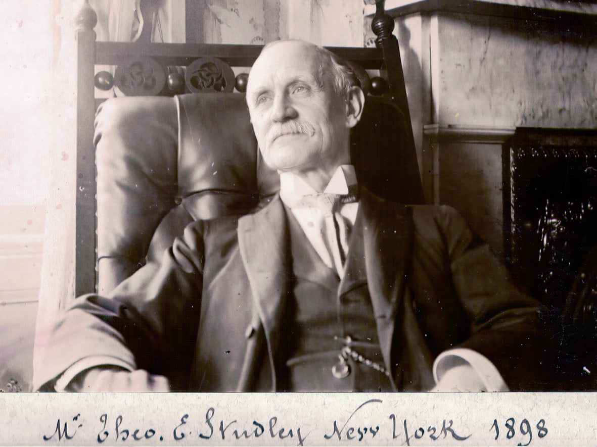 Photographie ancienne en noir et blanc d'un homme âgé en costume trois-pièces sombre, assis dans un fauteuil de cuir ; sous l'image, une légende manuscrite indique « M. Theo. E. Studley New York 1898 ».